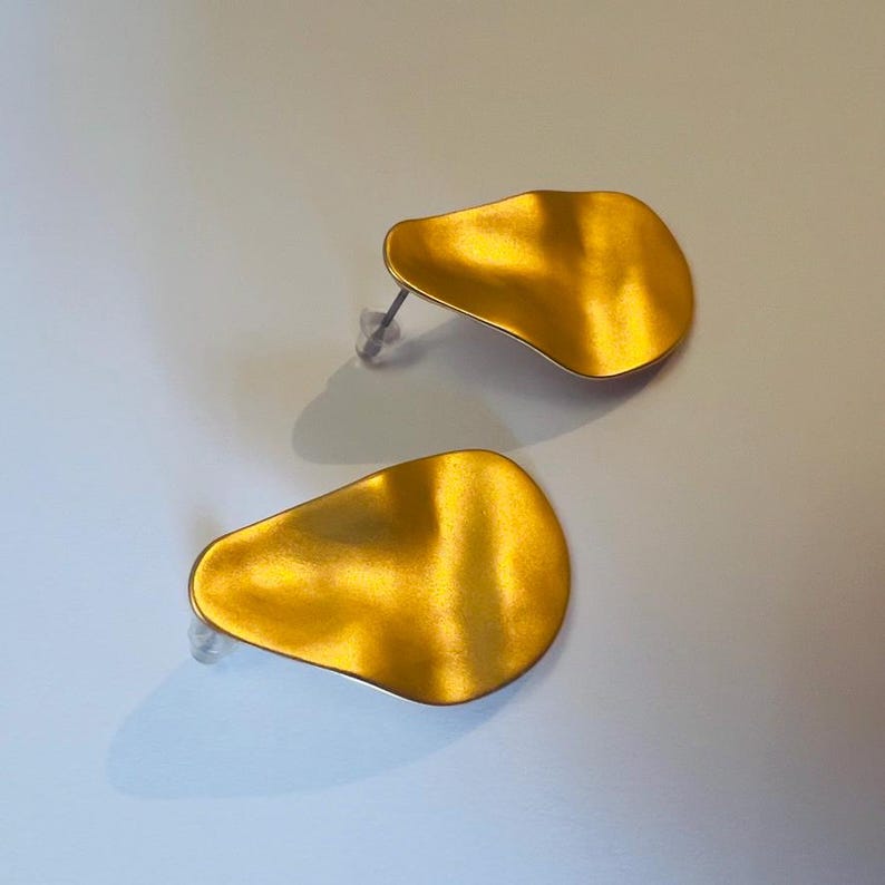 May include: A pair of gold-coloured stud earrings. Each earring has a unique, organic shape with a textured surface. The earrings are set against a light background, highlighting their metallic sheen and design.