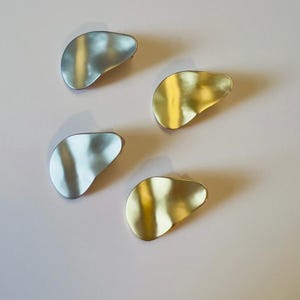 May include: Four abstract metal brooches in silver and gold tones are arranged on a light pink surface. The brooches have an organic, irregular shape, with a slightly textured surface. The silver brooches reflect light, while the gold ones have a warm, shiny appearance.