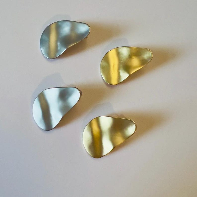 May include: Four abstract metal brooches in silver and gold tones are arranged on a light pink surface. The brooches have an organic, irregular shape, with a slightly textured surface. The silver brooches reflect light, while the gold ones have a warm, shiny appearance.