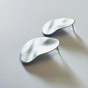 May include: A pair of silver stud earrings. Each earring has an irregular, organic shape with a rippled surface and a dark border. The earrings are displayed on a light grey surface, casting shadows.