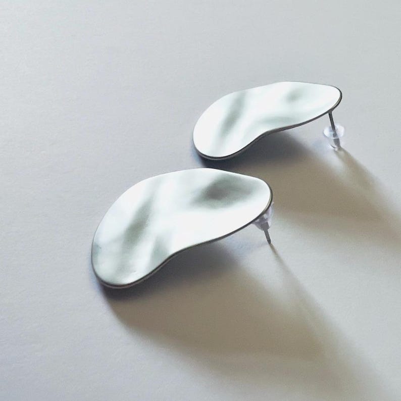 May include: A pair of silver stud earrings. Each earring has an irregular, organic shape with a rippled surface and a dark border. The earrings are displayed on a light grey surface, casting shadows.
