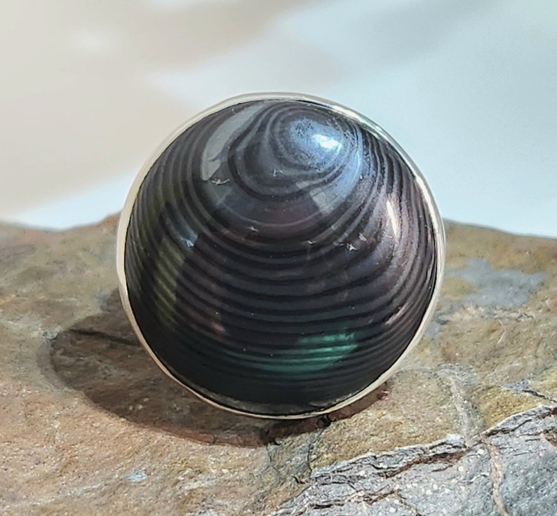 Puede incluir: Un anillo de plata con una piedra preciosa negra y verde. La piedra preciosa tiene un patr&oacute;n de remolino.