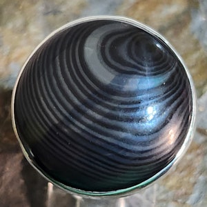 Puede incluir: Un anillo de plata con una piedra negra y gris con bandas. La piedra tiene un patr&oacute;n de remolino.