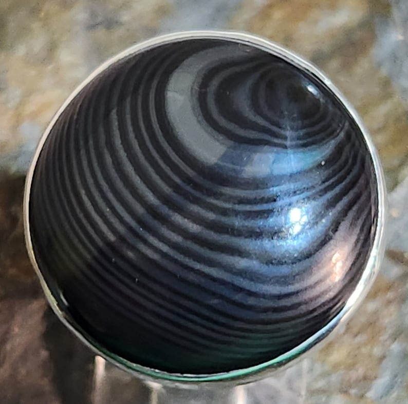 Puede incluir: Un anillo de plata con una piedra negra y gris con bandas. La piedra tiene un patr&oacute;n de remolino.