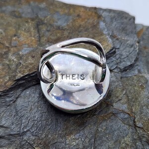 Puede incluir: Un anillo de plata con una piedra redonda verde engastada en el centro. El anillo est&aacute; marcado con "THEIS STERLING" en la parte posterior.