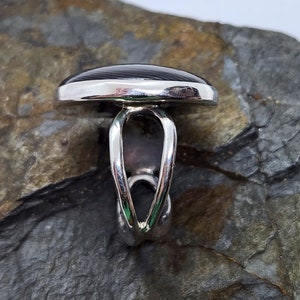 Puede incluir: Un anillo de plata con una piedra ovalada negra y gris. El anillo tiene una banda &uacute;nica y retorcida.