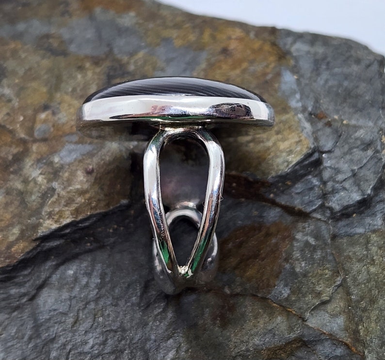 Puede incluir: Un anillo de plata con una piedra ovalada negra y gris. El anillo tiene una banda &uacute;nica y retorcida.