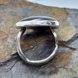 Puede incluir: Un anillo de plata con una piedra ovalada gris oscura engastada en el centro. El anillo est&aacute; sobre un fondo de roca gris y marr&oacute;n.