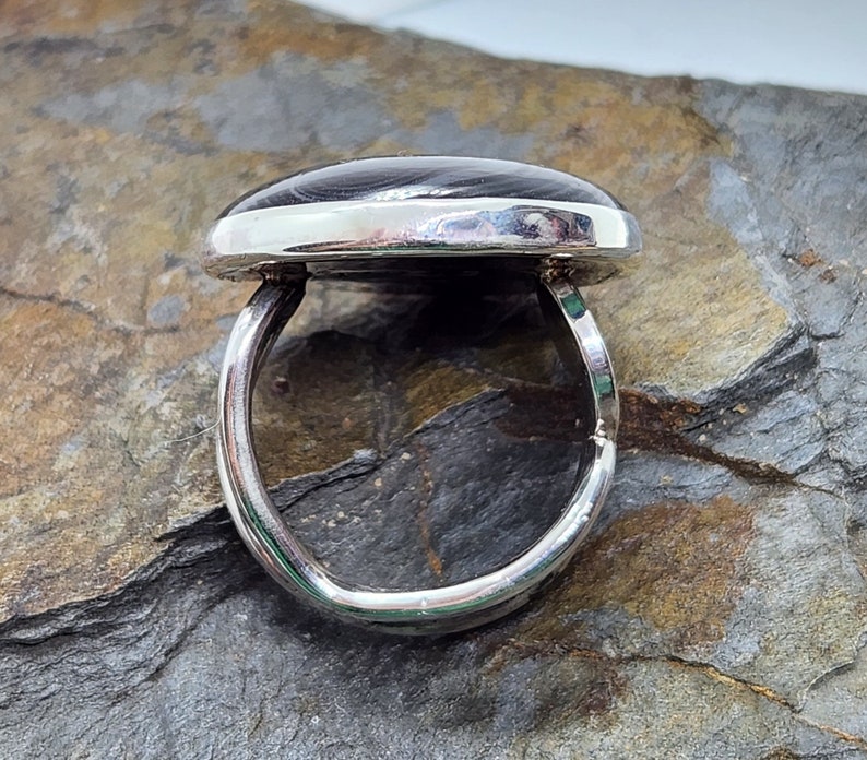 Puede incluir: Un anillo de plata con una piedra ovalada gris oscura engastada en el centro. El anillo est&aacute; sobre un fondo de roca gris y marr&oacute;n.