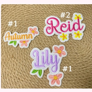 Puede incluir: Parches bordados con detalles florales. Los parches presentan los nombres "Autumn", "Reid" y "Lily" en varios colores. Cada parche tiene un diseño floral con detalles rosas, amarillos y verdes. Los parches están sobre un fondo texturizado y neutro.