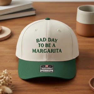 May include: A cream and green baseball cap with the text "BAD DAY TO BE A MARGARITA". The cap has a green peak and a small green button on top. The text is in green. The cap is a casual accessory.