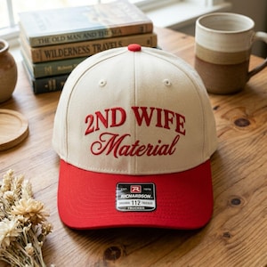 May include: A beige and red trucker hat with the embroidered text "2ND WIFE Material" in red. The hat has a curved brim and a Richardson tag. The hat is sitting on a wooden surface.