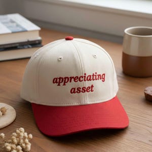 May include: A baseball cap with a cream-colored crown and a red brim. The cap features the embroidered text "appreciating asset" in red. The cap has a red button on top.