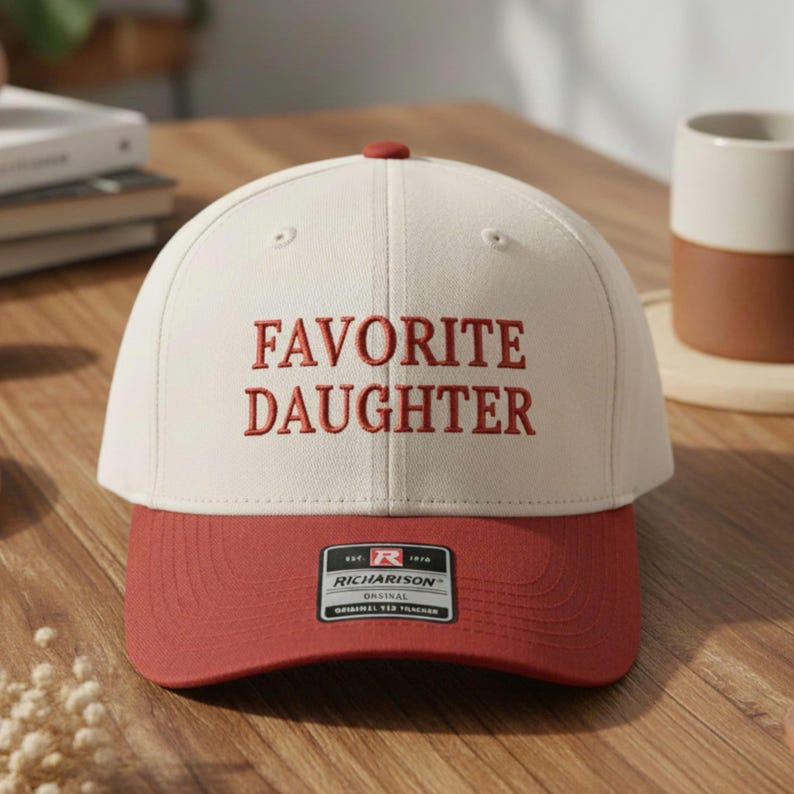 May include: A baseball cap with a white crown and a red brim. The words "FAVORITE DAUGHTER" are embroidered in red on the front. The cap has a curved brim and a small red button on top. A Richardson brand tag is visible.