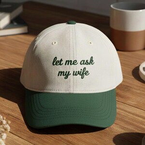 May include: A white and green baseball cap with the text "let me ask my wife" embroidered in green. The cap has a curved brim and a green button on top. The hat is sitting on a wooden surface.