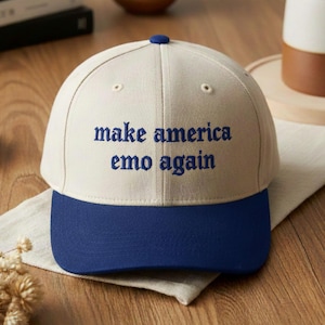 May include: A baseball cap with a cream-colored crown and a navy blue brim. The phrase "make america emo again" is embroidered in navy blue on the front of the cap. The cap has a navy blue button on top.