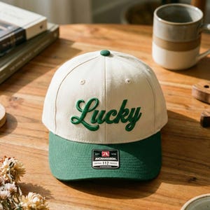 May include: Cream and green baseball cap featuring the word "Lucky" in green script. The cap has a green peak and a small green button on top. Includes a Richardson 112 trucker patch.