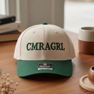 May include: A cream and green baseball cap with the embroidered text "CMRAGRL". The cap features a green peak and top button. A Richardson tag is visible on the front.