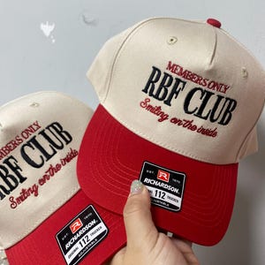 May include: Two beige and red baseball caps with the text "MEMBERS ONLY RBF CLUB Smiling on the inside" embroidered on the front. Each cap has a red brim and a Richardson 112 Trucker tag.