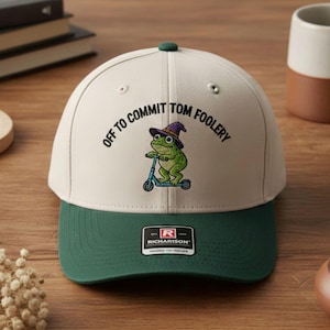 Op de afbeelding: Een crème en donkergroene baseball cap met de tekst "OFF TO COMMIT TOM FOOLERY" en een cartoon kikker met een heksenhoed op een scooter. De cap heeft een donkergroene klep en een groene knop.