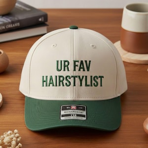 May include: A cream and green baseball cap with the text "UR FAV HAIRSTYLIST" embroidered in green. The cap has a green brim and a small green button on top. A black and white tag is visible on the front.