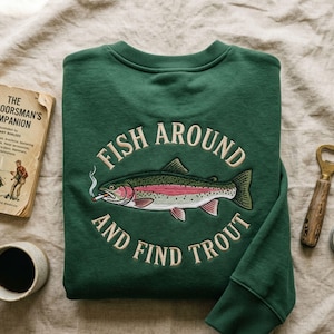 May include: A dark green sweatshirt featuring the embroidered text "FISH AROUND AND FIND TROUT" around a detailed trout illustration with a cigarette. The sweatshirt is folded on a neutral-toned surface.