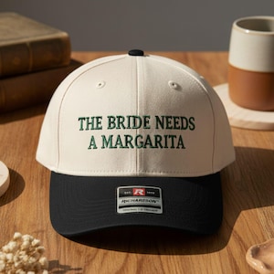 May include: A cream-colored baseball cap with a black brim. The text "THE BRIDE NEEDS A MARGARITA" is embroidered in green on the front. The cap has a black button on top and a Richardson label.