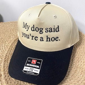 Puede incluir: Gorra de camionero beige y negra con el texto "My dog said you're a hoe." La gorra tiene una visera negra y un pequeño botón azul marino. La etiqueta de la marca Richardson es visible.
