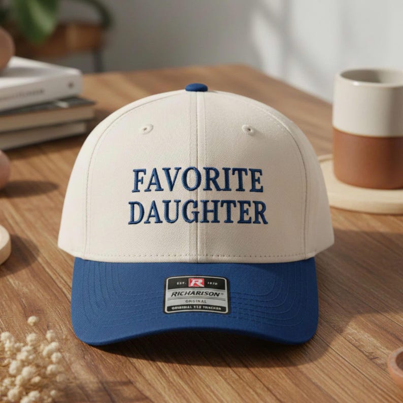 May include: A baseball cap with a cream crown and a blue peak. The words "FAVORITE DAUGHTER" are embroidered in blue on the front. The cap has a curved peak and a small blue button on top.