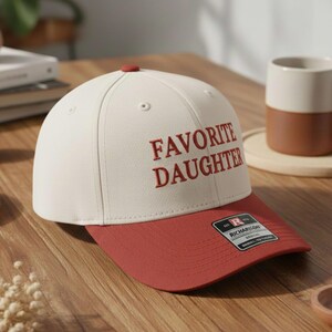 May include: A cream and red baseball cap with the embroidered text "FAVORITE DAUGHTER". The cap has a red peak and button, and a Richardson brand tag. The hat is on a wooden surface.