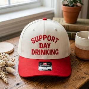 May include: A cream-colored baseball cap with a red brim and top button. The words "SUPPORT DAY DRINKING" are embroidered in red across the front. A black and white tag is on the front of the cap.