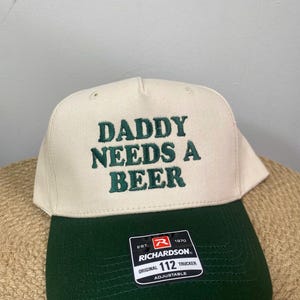 May include: A beige and green trucker hat with the text "DADDY NEEDS A BEER" embroidered in green. The hat has a curved brim and an adjustable strap. The Richardson 112 Trucker hat is a classic style.