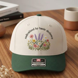 Op de afbeelding: Crèmekleurige baseballcap met een groene klep en knoop. De cap heeft een geborduurd bloemendesign en de tekst "radicalized by basic decency". Een Richardson-merklabel is zichtbaar.