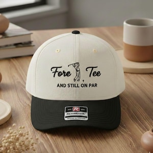May include: A cream-colored baseball cap with a black brim. The cap features the text "Fore Tee" with a golf-themed illustration and the phrase "AND STILL ON PAR." A small "Richardson" patch is visible on the front.