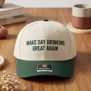 May include: A cream-coloured baseball cap with a green peak and top button. The cap has the embroidered phrase "MAKE DAY DRINKING GREAT AGAIN" in green lettering. A black and white Richardson label is on the front.