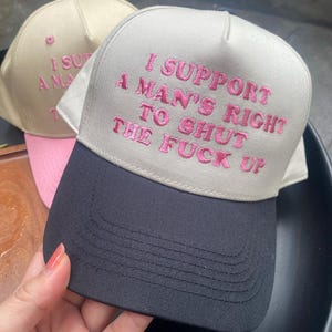 May include: Two baseball caps. One is tan with a pink brim and pink embroidered text. The other is gray with a black brim and pink embroidered text that reads "I SUPPORT A MAN'S RIGHT TO SHUT THE FUCK UP."