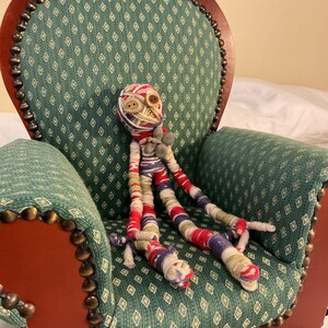 May include: A handmade doll with button eyes and wrapped yarn head and limbs, seated in a small antique chair. The chair has a green patterned fabric back and seat, with a red wooden frame and brass nailhead trim.