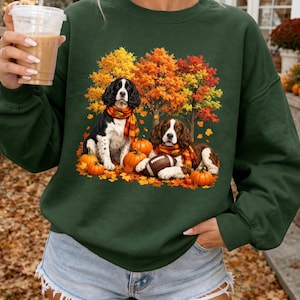 May include: A dark green crewneck sweatshirt featuring a graphic print of two spaniel dogs wearing plaid scarves, sitting among orange pumpkins and autumn leaves with trees in the background. One dog is resting its paw on a brown football.