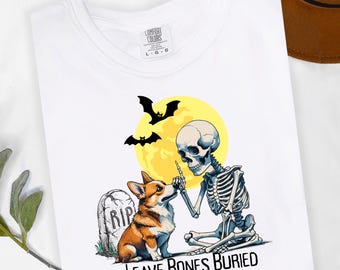 May include: A white t-shirt featuring a graphic of a skeleton sitting next to a Corgi dog in front of a tombstone marked RIP. Above them is a yellow full moon and two black bats. The text "LEAVE BONES BURIED" is printed in black at the bottom.