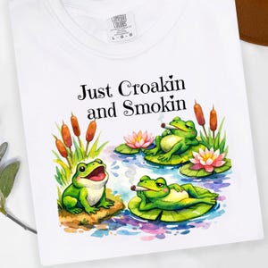 May include: White t-shirt featuring a watercolor illustration of three green frogs on lily pads, with cattails and pink water lilies. The frogs are smoking and the text "Just Croakin and Smokin" is above the illustration.
