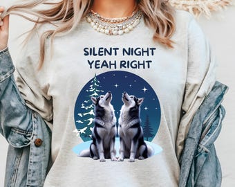 May include: A light-colored crewneck sweatshirt featuring the dark blue text "SILENT NIGHT YEAH RIGHT" above a graphic of two grey and white huskies sitting in a snowy forest under a dark blue night sky with stars and a pine tree silhouette.