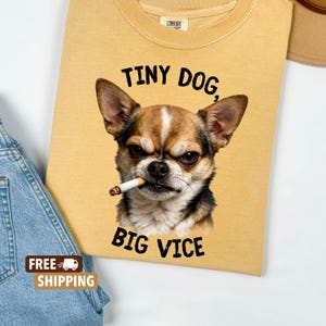 May include: Mustard yellow sweatshirt featuring a chihuahua with a cigarette in its mouth. The text "TINY DOG, BIG VICE" is printed above and below the dog. The sweatshirt has a crew neck and a small tag at the top.