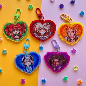 May include: Five heart-shaped keyrings featuring anime-style character illustrations. Each keyring showcases a different character with unique hair and clothing, surrounded by a coloured border. The keyrings have colourful clasps and star-shaped decorations.