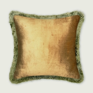 May include: A square throw pillow featuring a metallic gold, textured fabric center panel. The pillow is bordered by a thick, sage green fringe trim that extends along all four edges, creating a distinct contrast with the shimmering gold surface.