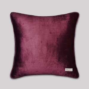 May include: A square, plum-colored velvet pillow with a decorative rope trim. The pillow has a soft, textured appearance and a small rectangular label in the lower right corner with the word "Prestige" printed on it.