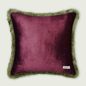 May include: A square decorative pillow with a deep burgundy velvet center and a sage green fringe border. The pillow has a small white tag with the word "Predier" on it. The pillow is a home decor item.