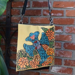 May include: A yellow canvas tote bag with a black strap and a hand-painted design of blue, orange, and green flowers. The bag is signed "Juliana Martinez".
