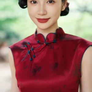 May include: A woman in a sleeveless, red satin dress with a mandarin collar and teal trim. The dress features a delicate floral pattern. Her dark hair is styled with a side part and a small flower.