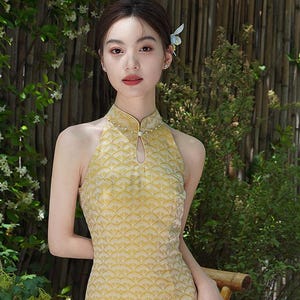 May include: A yellow halter-neck dress with a repeating scale pattern. The dress has a keyhole neckline and is adorned with small pearls. The dress is form-fitting and reaches the ankles. The background features greenery and a bamboo structure.