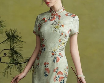 May include: A light green, short-sleeved, floral-print dress with a mandarin collar. The dress features red and yellow flowers and a side slit. The woman is holding a fan and wearing a flower in her hair.